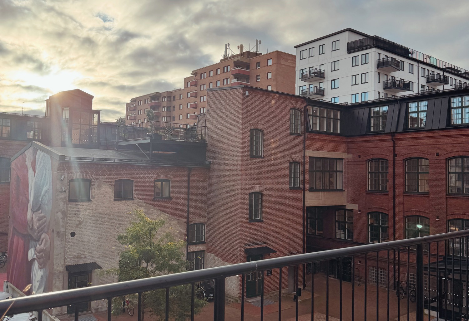 En innergård i ett äldre industriområde med röda tegelbyggnader och moderna bostadshus i bakgrunden, sedd från en balkong med svart räcke. Till vänster syns en stor väggmålning av en person som håller ett barn, några parkerade bilar och ett träd planterat i en rund betonglåda. Himlen är molnig med morgon- eller kvällssol som lyser in från vänster. Trikåfabrikens innergård Malmö