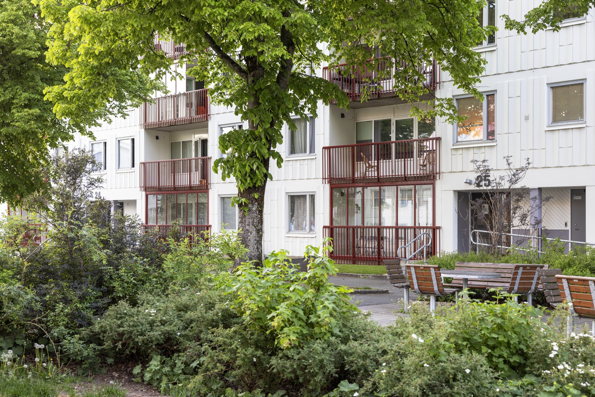 Ett flerfamiljshus i vitt med röda balkonger. Framför byggnaden finns grönskande buskar och träd samt några parkbänkar.