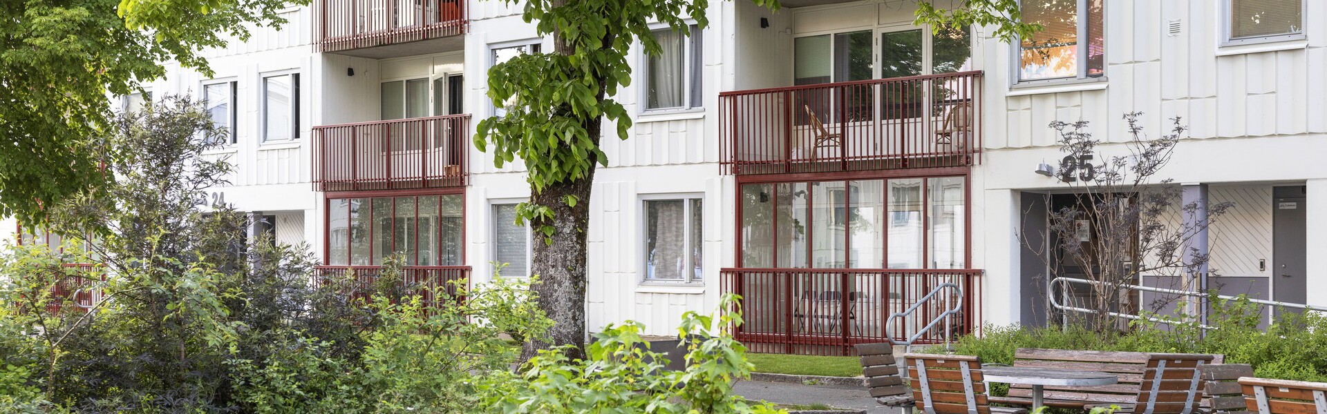 Ett flerfamiljshus i vitt med röda balkonger. Framför byggnaden finns grönskande buskar och träd samt några parkbänkar.