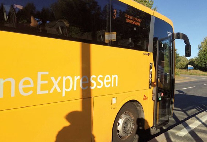 En gul buss från SkåneExpressen står stilla vid en hållplats en solig dag. Bussens dörr är öppen och en skugga från en person syns på sidan av bussen. Destinationen på skylten visar 'Simrishamn'.