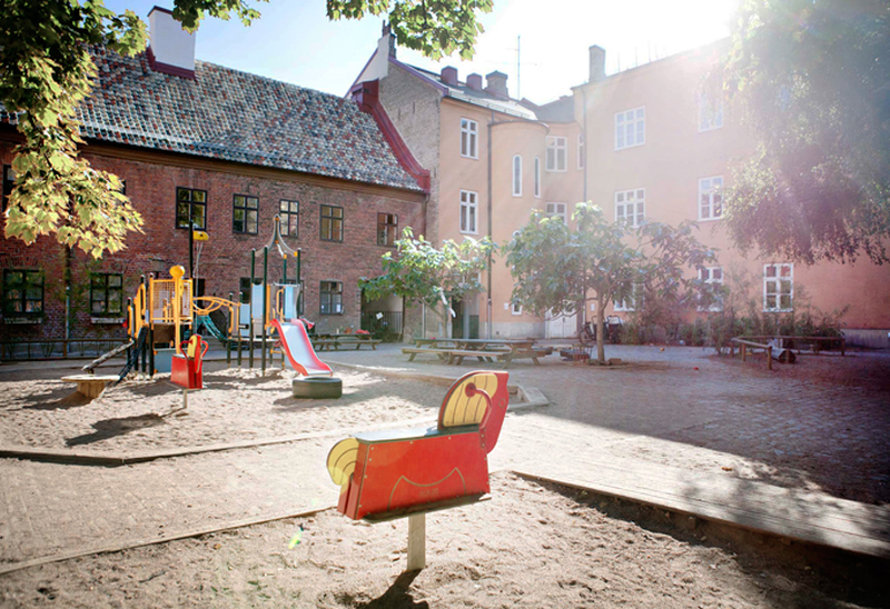 En solbelyst innergård med en lekplats som innehåller rutschkana, klätterställning och fjädergungor i färgglada färger. Gården är omgiven av äldre flerbostadshus i tegel med stora fönster. Träd och skuggor från lövverket ramar in platsen, och solens strålar skapar en varm, inbjudande atmosfär.