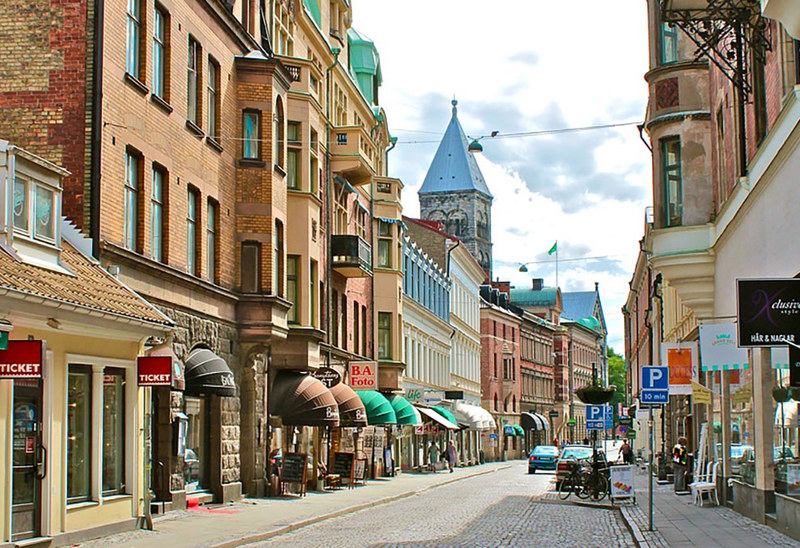 Stadsgata i centrala Lund med äldre byggnader i olika stilar längs båda sidor. Flera butiker och restauranger med skyltar och markiser syns i gatubilden. Längst bort i bakgrunden reser sig Lunds domkyrka med sina karakteristiska torn. Några bilar är parkerade längs gatan och en cykel står lutad mot en stolpe