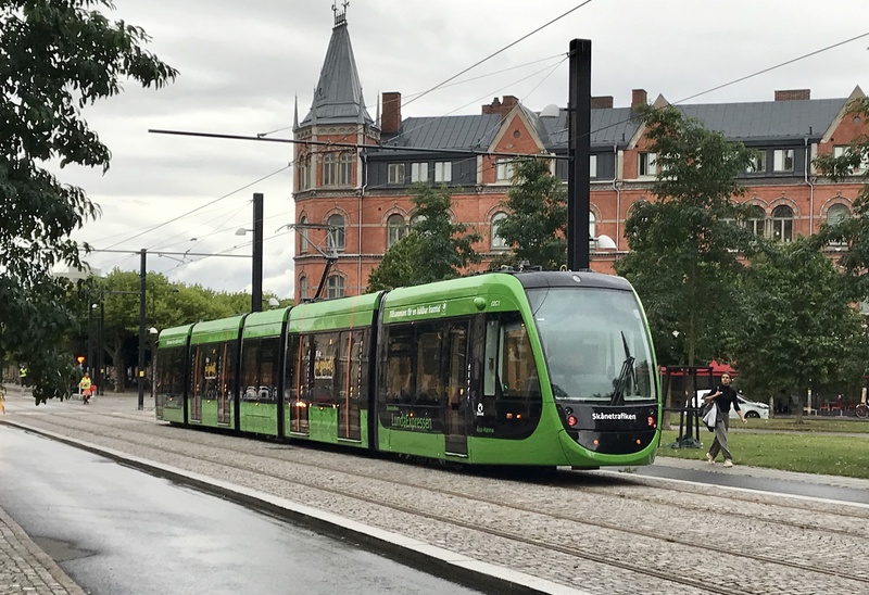 Grön spårvagn från Skånetrafiken på spårvägen i Lund, Sverige. Spårvagnen kör längs en gata med kullerstenar, omgiven av träd och ett stort rött tegelhus med torn i bakgrunden. Himlen är mulen och en person syns gå över gatan till höger.