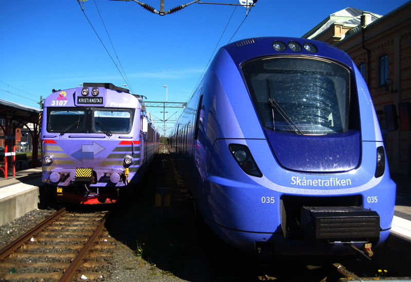 Två tåg står sida vid sida vid en perrong. Till vänster syns ett äldre, lila tåg med destinationen "Kristianstad". Till höger står ett modernt blått Pågatåg från Skånetrafiken med numret 035. Himlen är klarblå och en äldre stationsbyggnad syns i bakgrunden.
