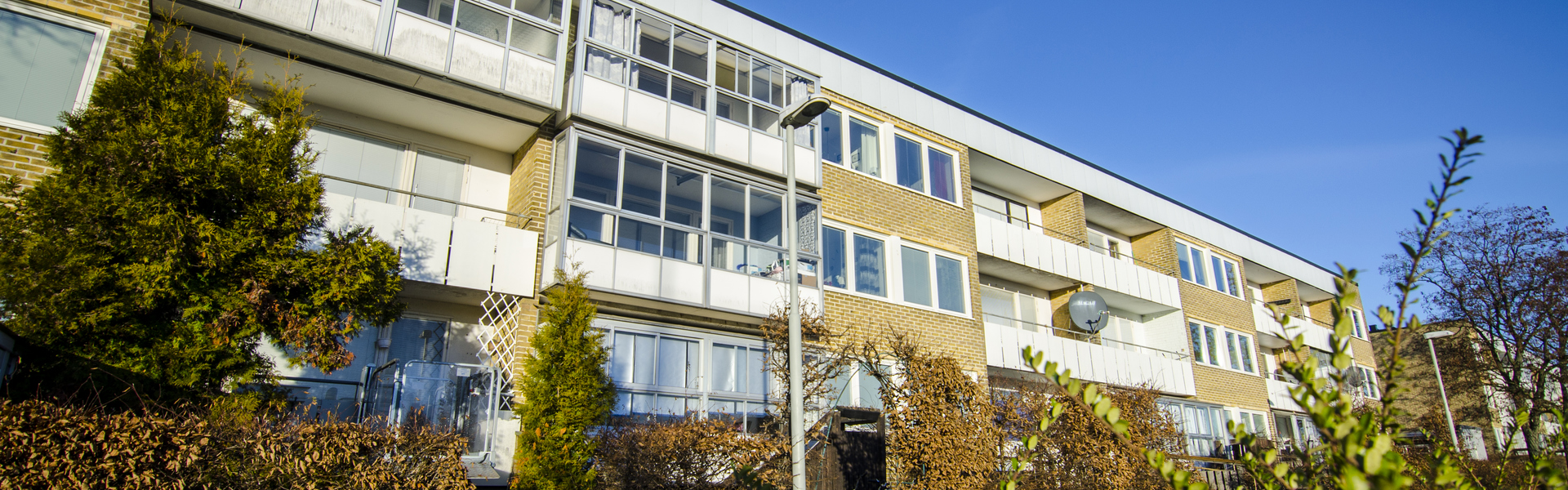 Ett trevånings flerbostadshus i gult tegel med stora inglasade balkonger. Framför huset växer välskötta buskar och träd, och en blå himmel breder ut sig ovanför byggnaden.