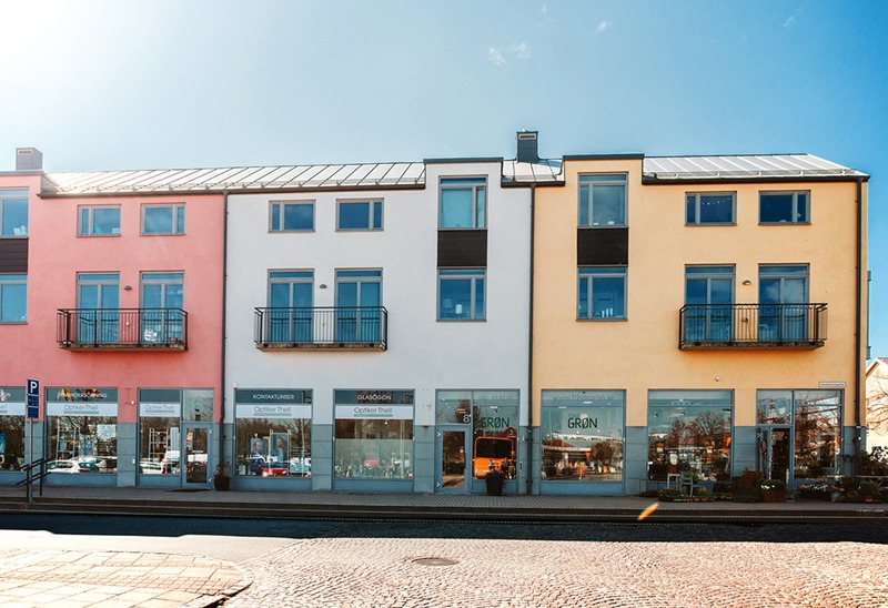 Flerbostadshus med butiker i bottenplan och färgglada fasader i rosa, vitt och gult. Husen har stora skyltfönster på gatuplan och små franska balkonger på de övre våningarna. Framför byggnaden syns en trottoar och en stenlagd gata i solljus.