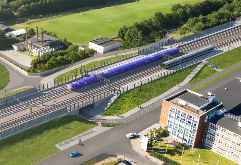 Ett pågatåg passerar genom ett grönområde nära en stad. Tåget kör på en upphöjd järnväg bredvid ett modernt kontorshus. I bakgrunden syns fotbollsplaner, flerbostadshus och mycket grönska.