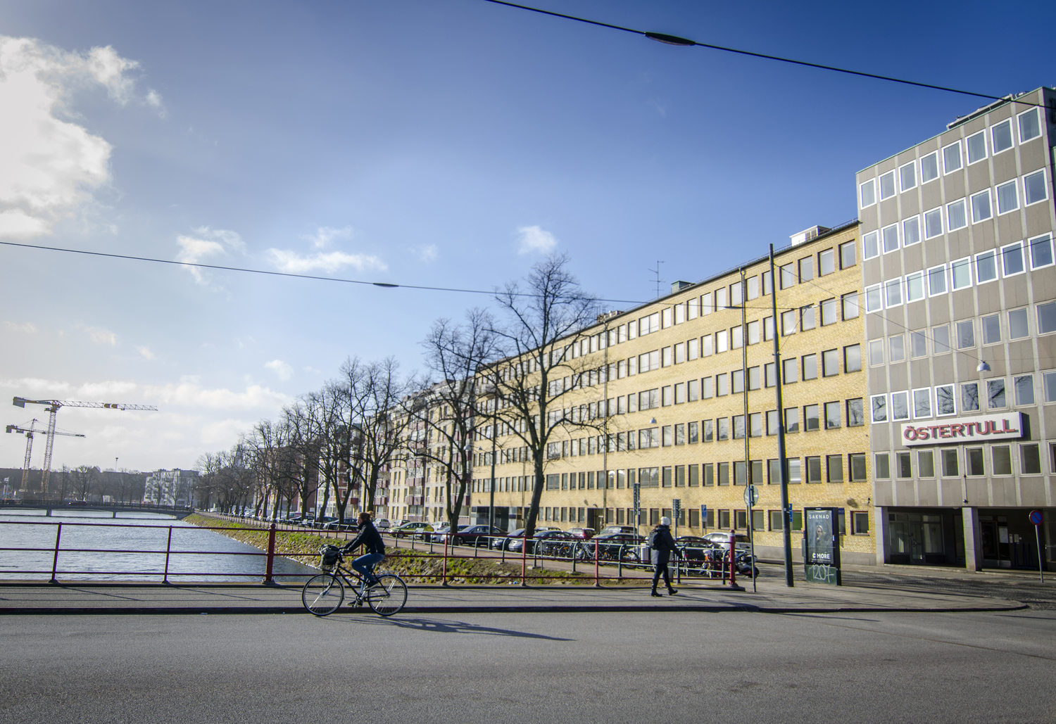 En stadsvy med en bred gata i förgrunden där en cyklist och en fotgängare rör sig. Till höger syns en gul och vit kontors- eller bostadsbyggnad med skylten "ÖSTERTULL" på fasaden. Till vänster om byggnaden sträcker sig en rad med träd längs ett vattendrag, och flera bilar står parkerade. I bakgrunden syns en bro och två byggkranar. Himlen är blå med några moln, och solen skiner.