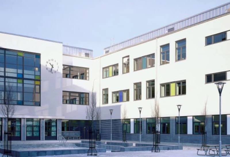 En modern skolbyggnad med vit fasad och stora fönsterpartier i varierande färger. Byggnaden omger en öppen innergård med bänkar, lampstolpar och unga träd. En stor klocka är synlig på ena väggen, och gården är stenbelagd med geometriska mönster.