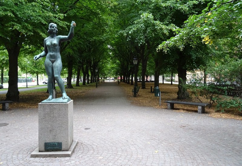 En staty av en naken kvinna i brons står på ett stenfundament i en allé med träd som bildar en tunnel över en gångväg. På båda sidor om gången finns parkbänkar och träd med gröna blad. Marken är täckt av stenplattor och vissna löv.