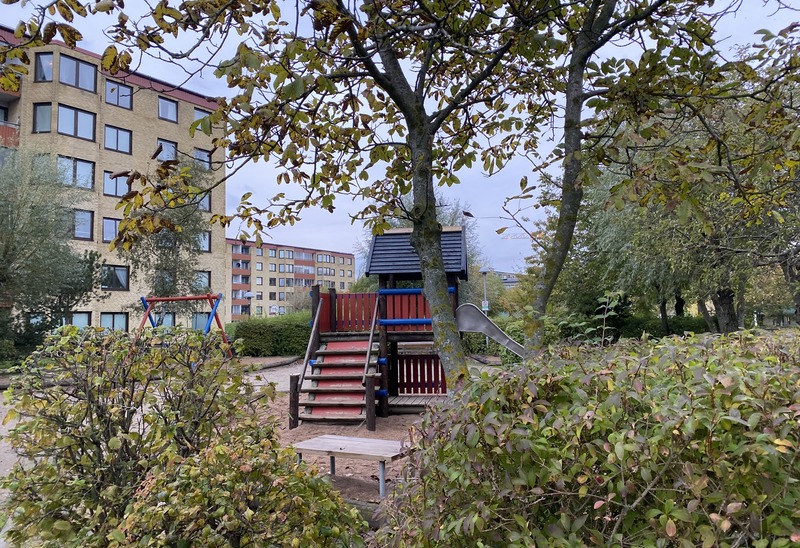 En lekplats med en trälekstruktur som har en rutschkana och trappor, omgiven av buskar och träd. I bakgrunden syns flerbostadshus med färgglada fasader. Höstlöv syns i träden och på marken.