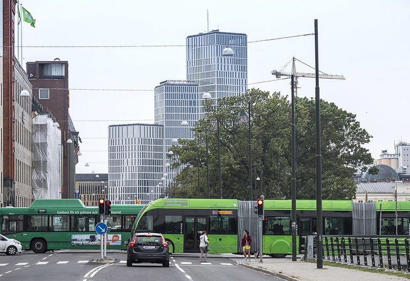 En grön buss korsar en gata i en stadsmiljö med moderna höghus och en byggkran i bakgrunden.
