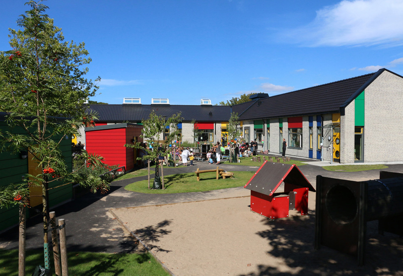 Förskolegård med flera barn som leker utomhus en solig dag. Gården omges av moderna byggnader med färgglada detaljer. Det finns lekstugor, träd och bänkar på området.
