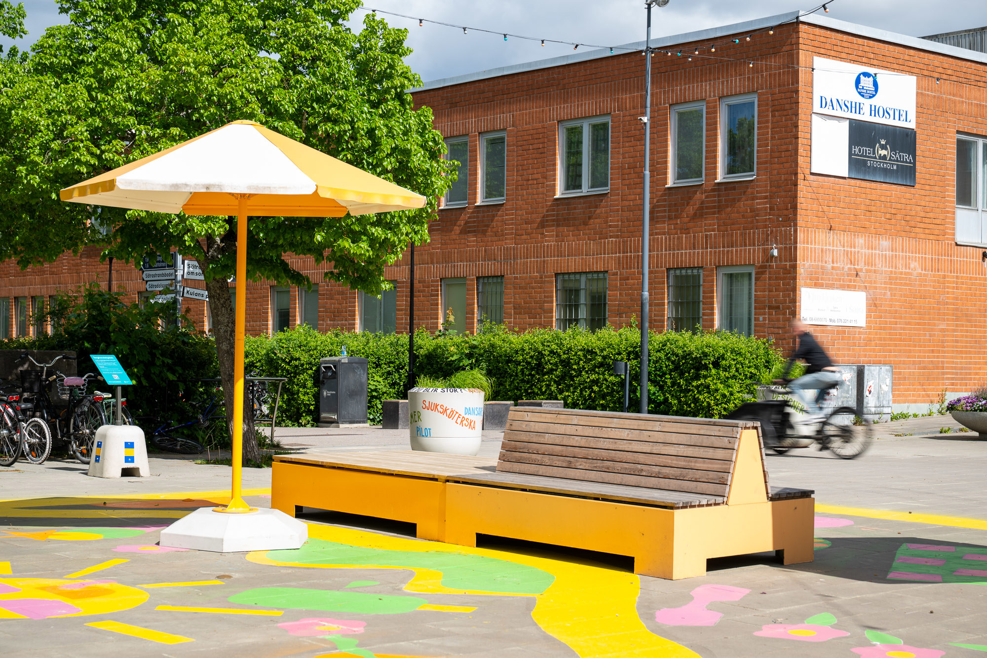 Tegelbyggnad i bakgrund med mötesplats med gul bänk och gult parasol i förgrund. 