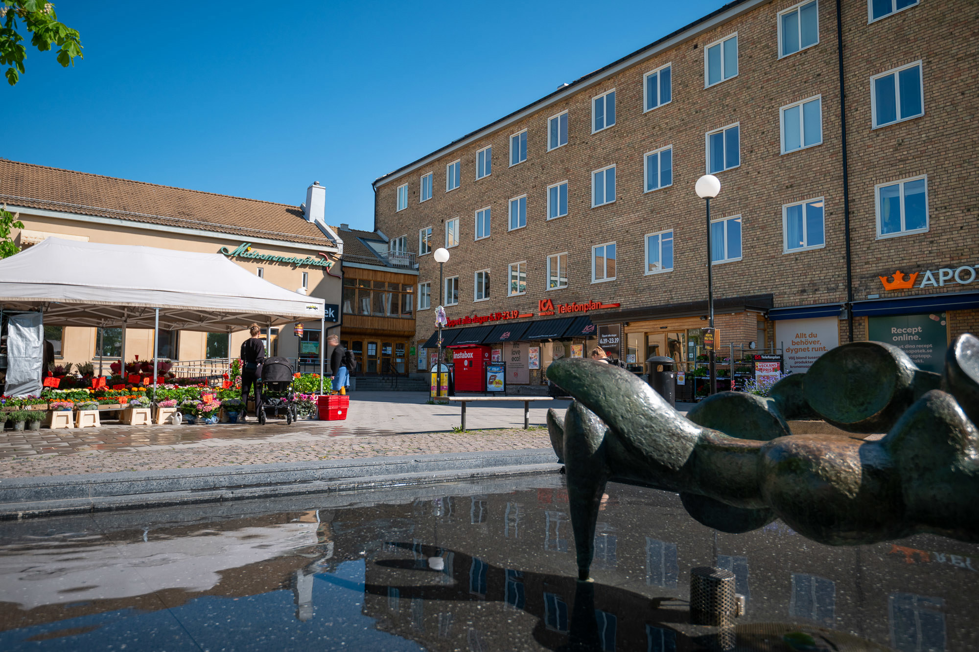Centrum torg omgicen av tegelbyggnader. Ett fruktstånd och en skulptur i vattendamm i förgrund