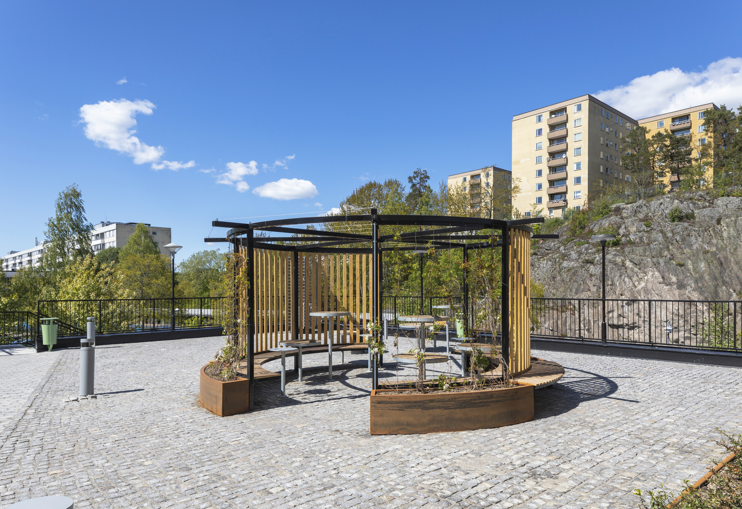 Plats med bänkar och bord omgiven av en rundad träbebyggelse på kullersten.