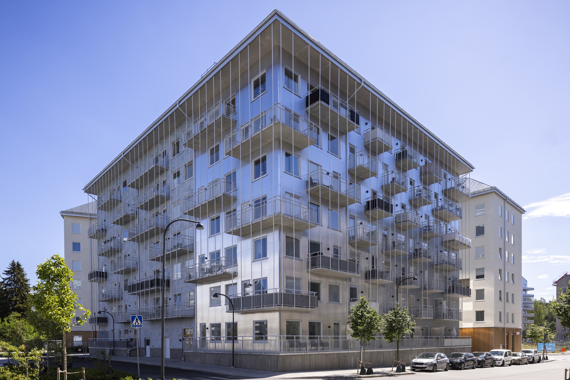 Flerbostadshus i modern stil med fasad i silverfärgad plåt och många utskjutande balkonger, sett från gatan en solig dag med blå himmel, trädplanteringar längs trottoaren och parkerade bilar framför huset. Supernova i Vega Haninge