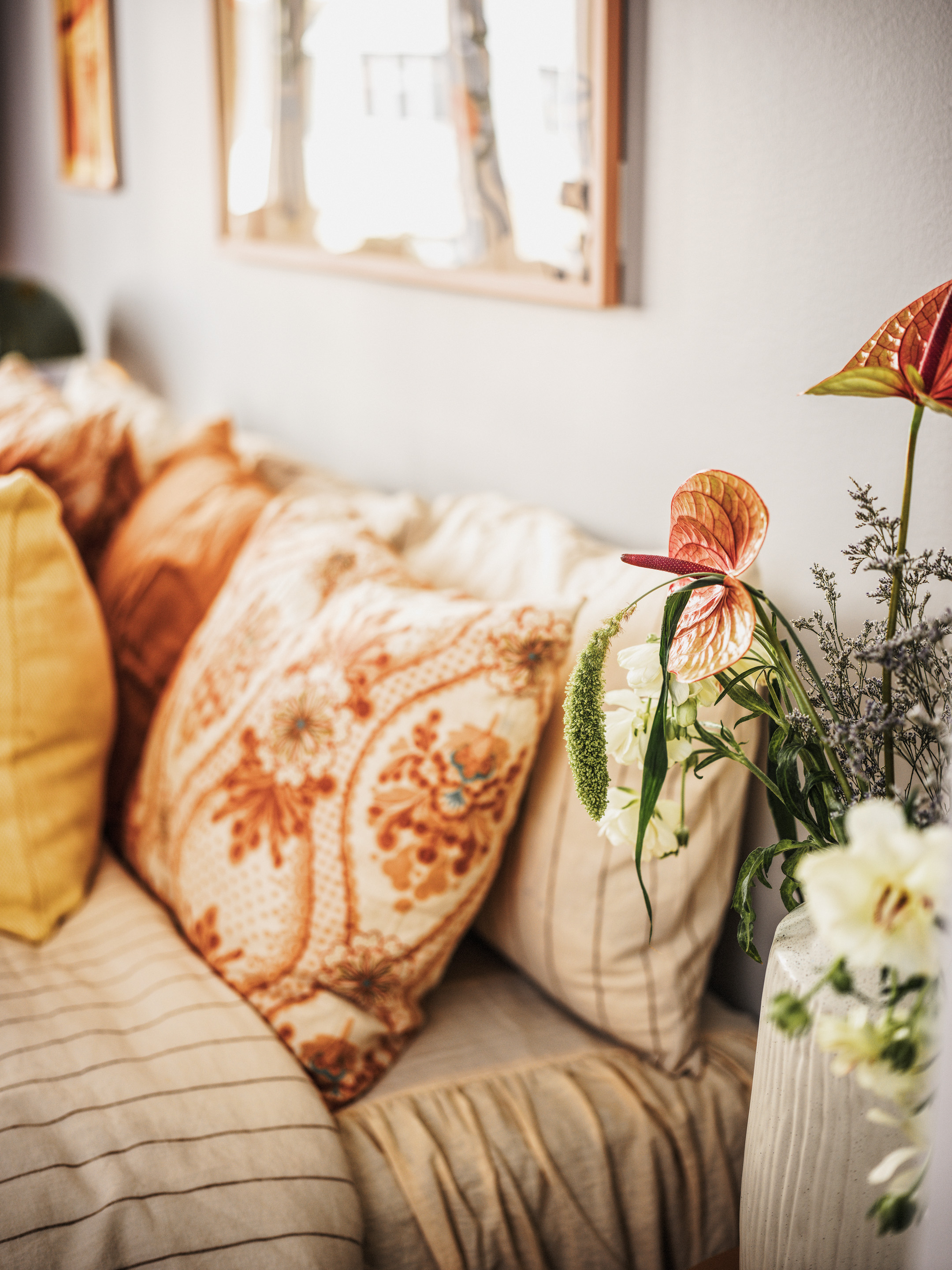 Detaljbild med hemtrevligt inredd del av rum med färska blommor i förgrunden och flera kuddar lutade mot en sänggavel