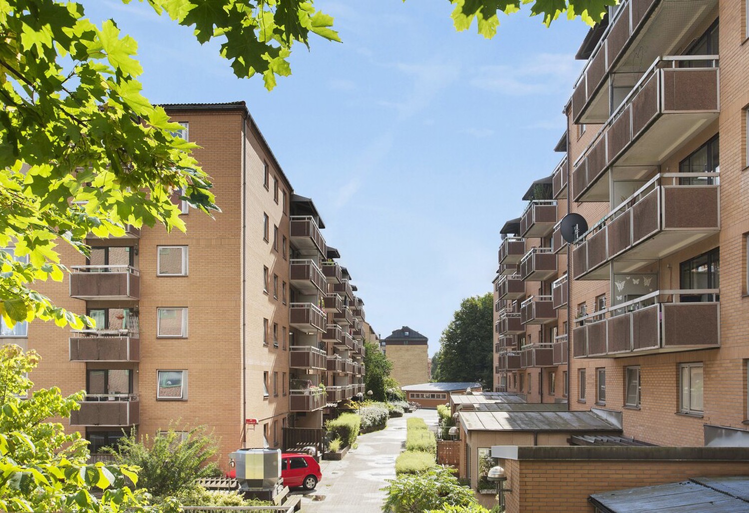 Vy mellan två rader tegelklädda flerbostadshus med balkonger, en grönskad gångväg emellan och träd i förgrunden under klarblå himmel.