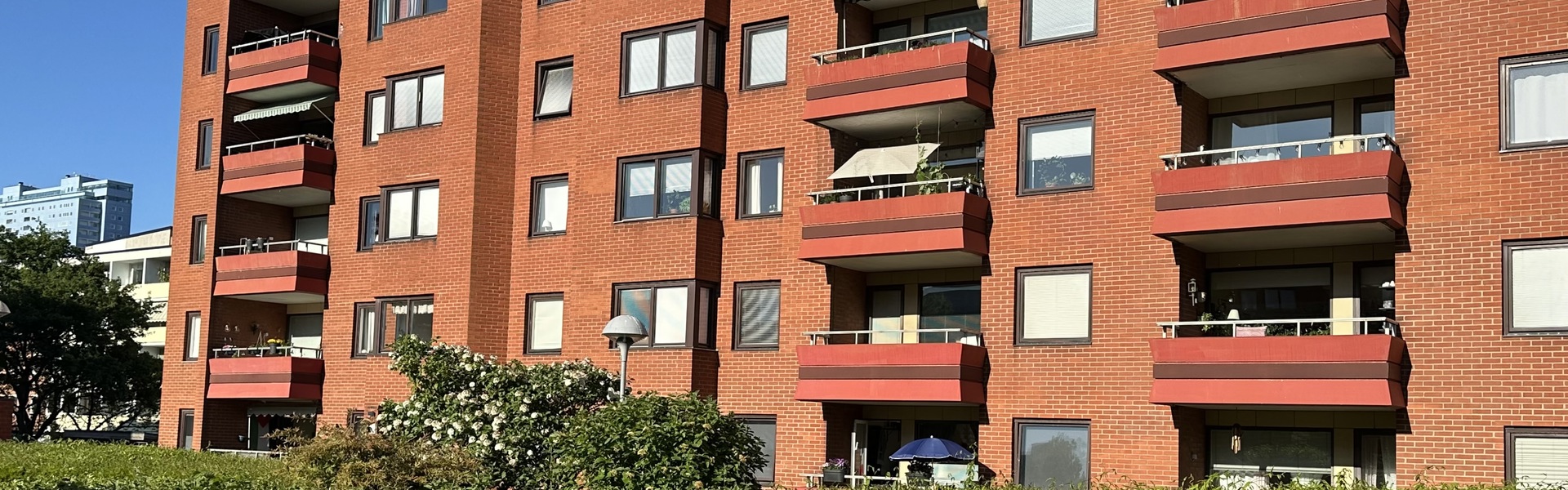 Ett rött flerbostadshus med flera våningar och balkonger i solsken, framför en grön häck och en innergård med en rund blomplantering i mitten.