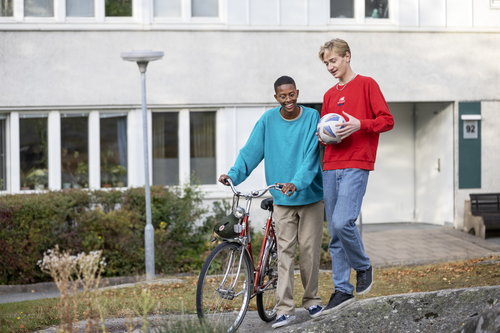 Två personer går tillsammans utomhus framför ett bostadshus. Den ena leder en cykel och den andra håller i en boll.