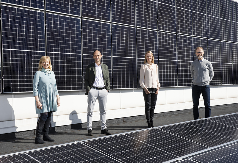 Fyra personer står på ett tak framför en rad solpaneler. De är klädda i kontorskläder och ser mot kameran. Solpanelerna täcker väggen bakom dem och en del av taket. Stämningen är professionell och fokuserad på hållbarhet.