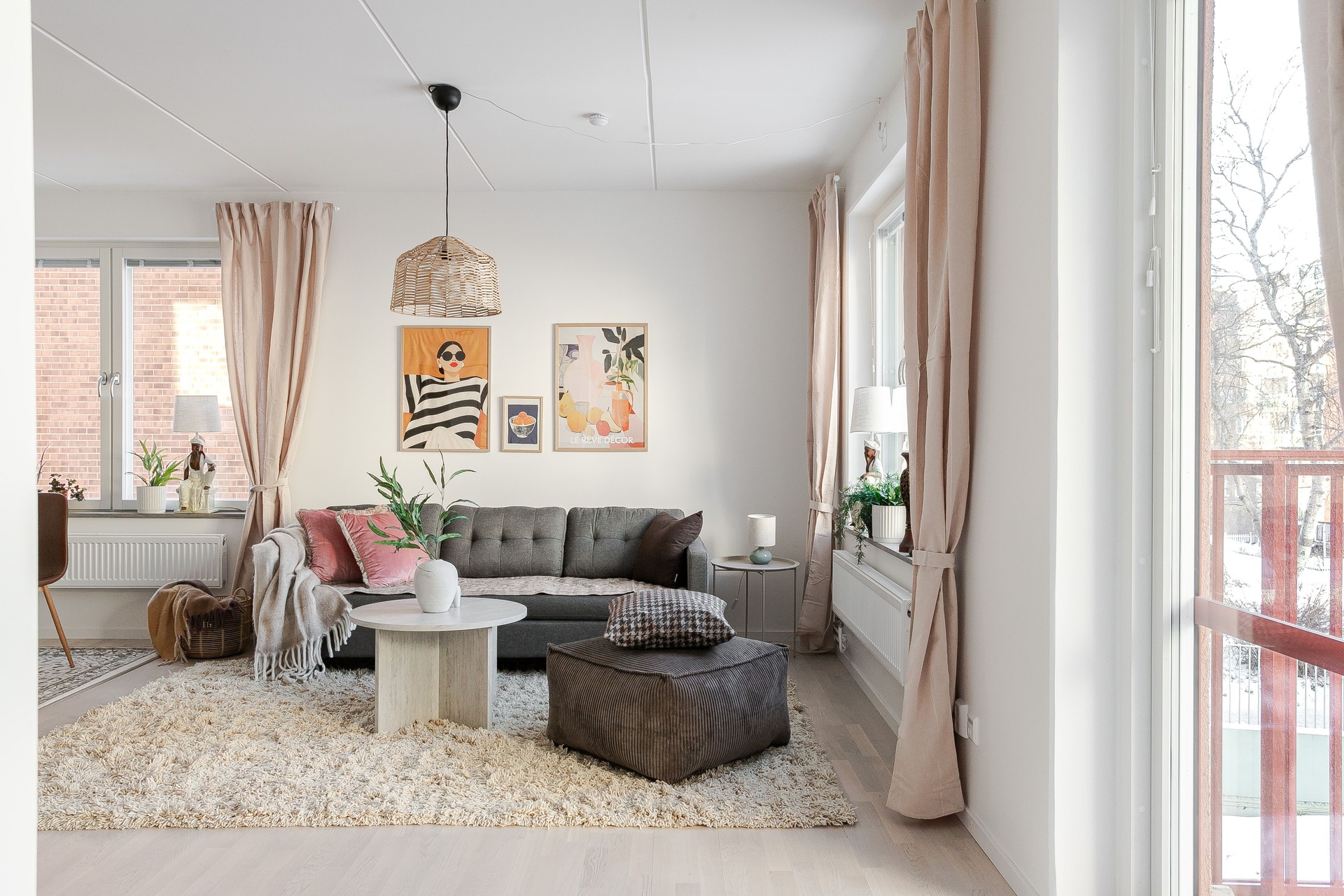 Ljust vardagsrum med grå soffa, brun sittpuff och runt soffbord på ljus ryamatta, stora fönster med beige gardiner samt tavlor och gröna växter som dekoration.