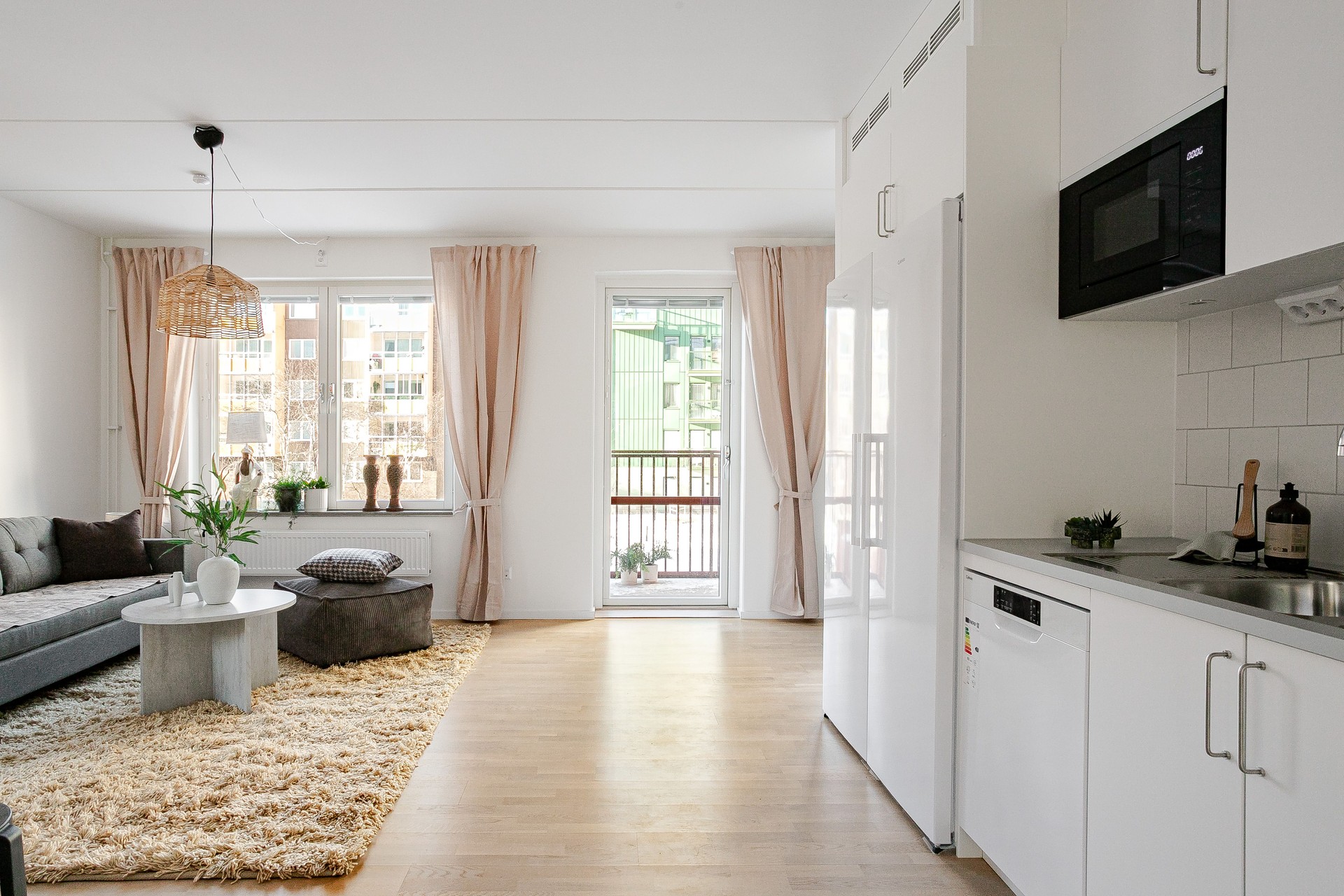 Ljust vardagsrum med öppen planlösning mot vit köksdel, grå soffa och soffbord på ljus ryamatta samt stora fönster och balkongdörr med beige gardiner.
