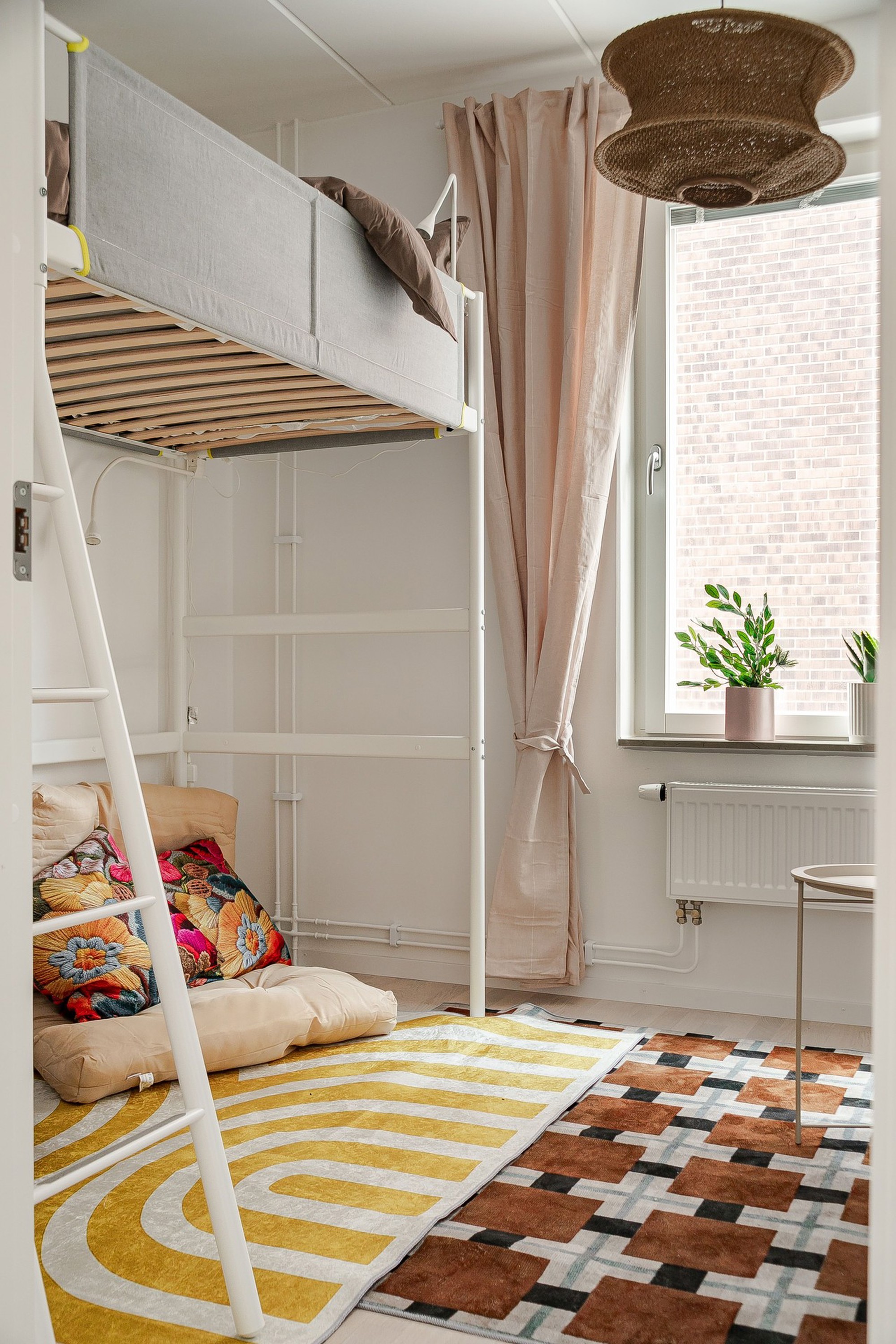 Barnrum med loftsäng, sittplats på golvet med kuddar, färgglada mönstrade mattor samt fönster med beige gardin och krukväxter.