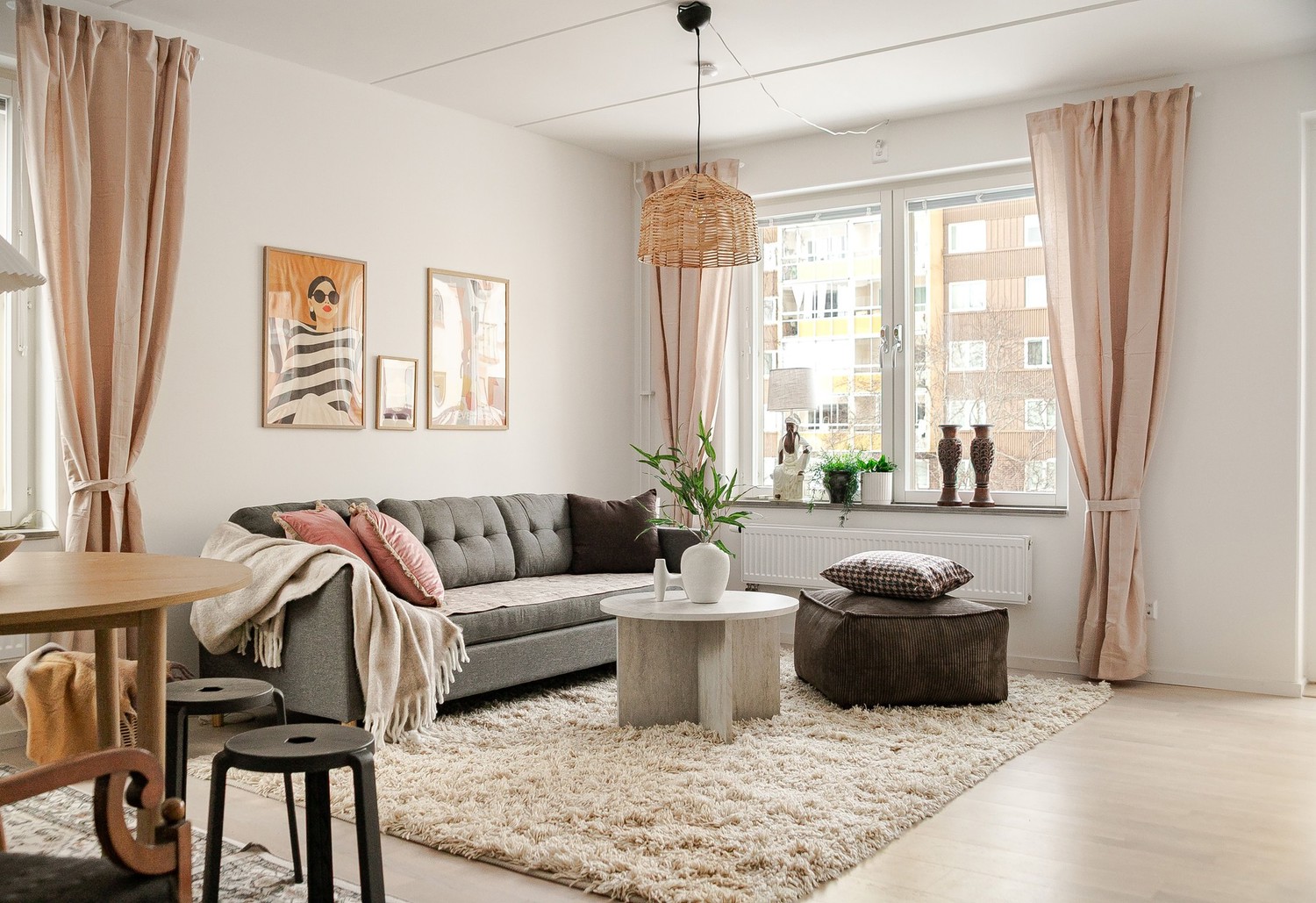 Ljust vardagsrum med grå soffa, runt soffbord och brun sittpuff på ljus ryamatta, stora fönster med beige gardiner samt tavlor och gröna växter som dekoration.