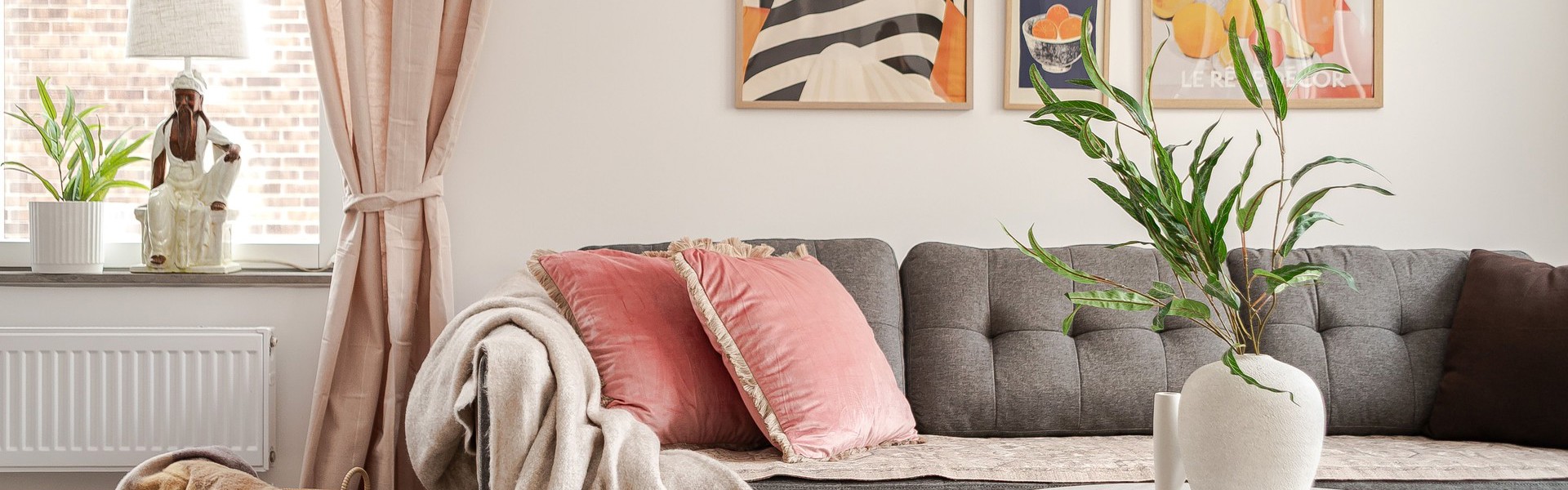 Ljust vardagsrum med grå soffa, rosa prydnadskuddar och pläd, runt soffbord med vit vas och grön växt, samt fönster med beige gardiner och färgstarka tavlor på väggen.