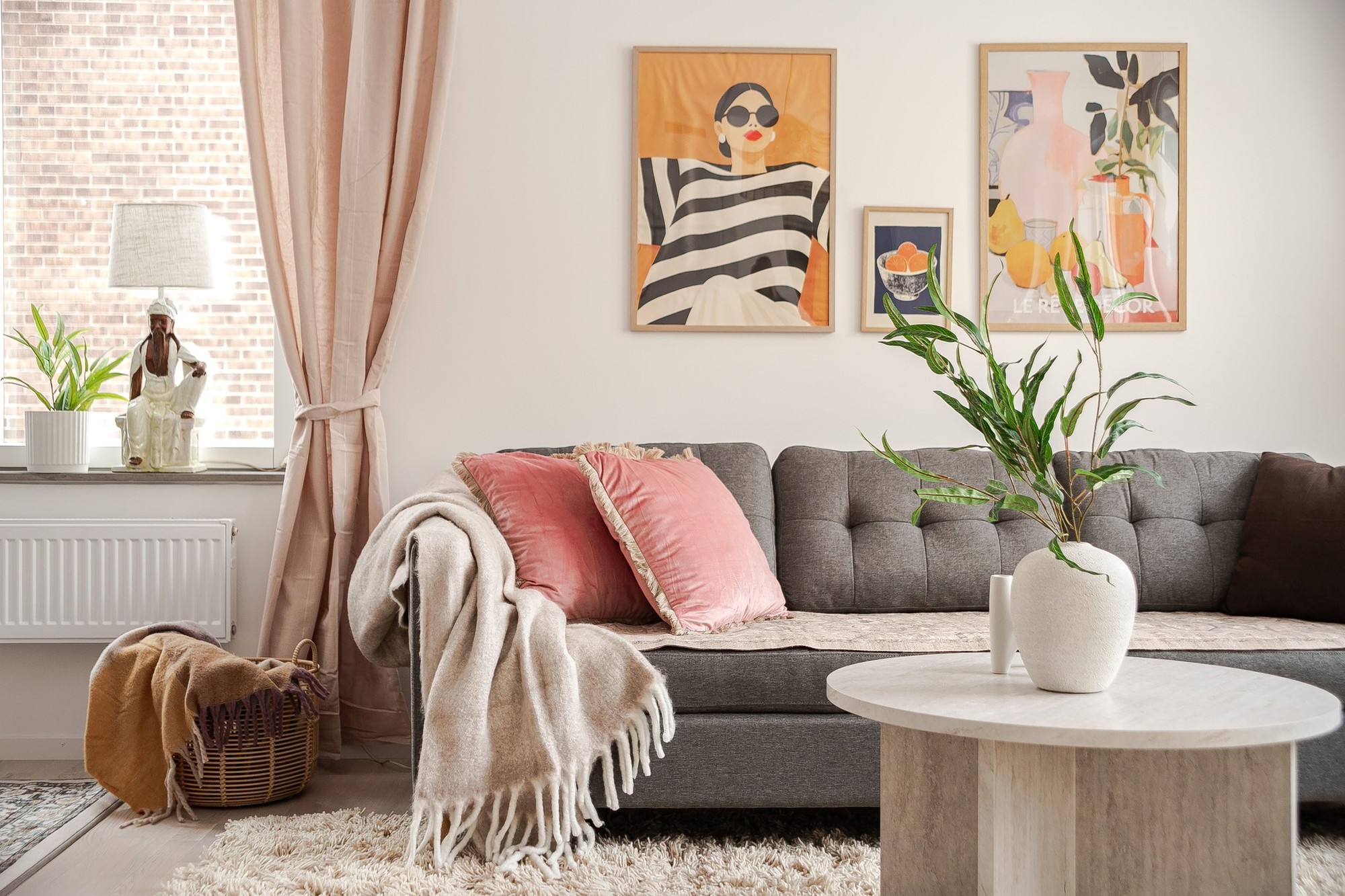 Ljust vardagsrum med grå soffa, rosa prydnadskuddar och pläd, runt soffbord med vit vas och grön växt, samt fönster med beige gardiner och färgstarka tavlor på väggen.
