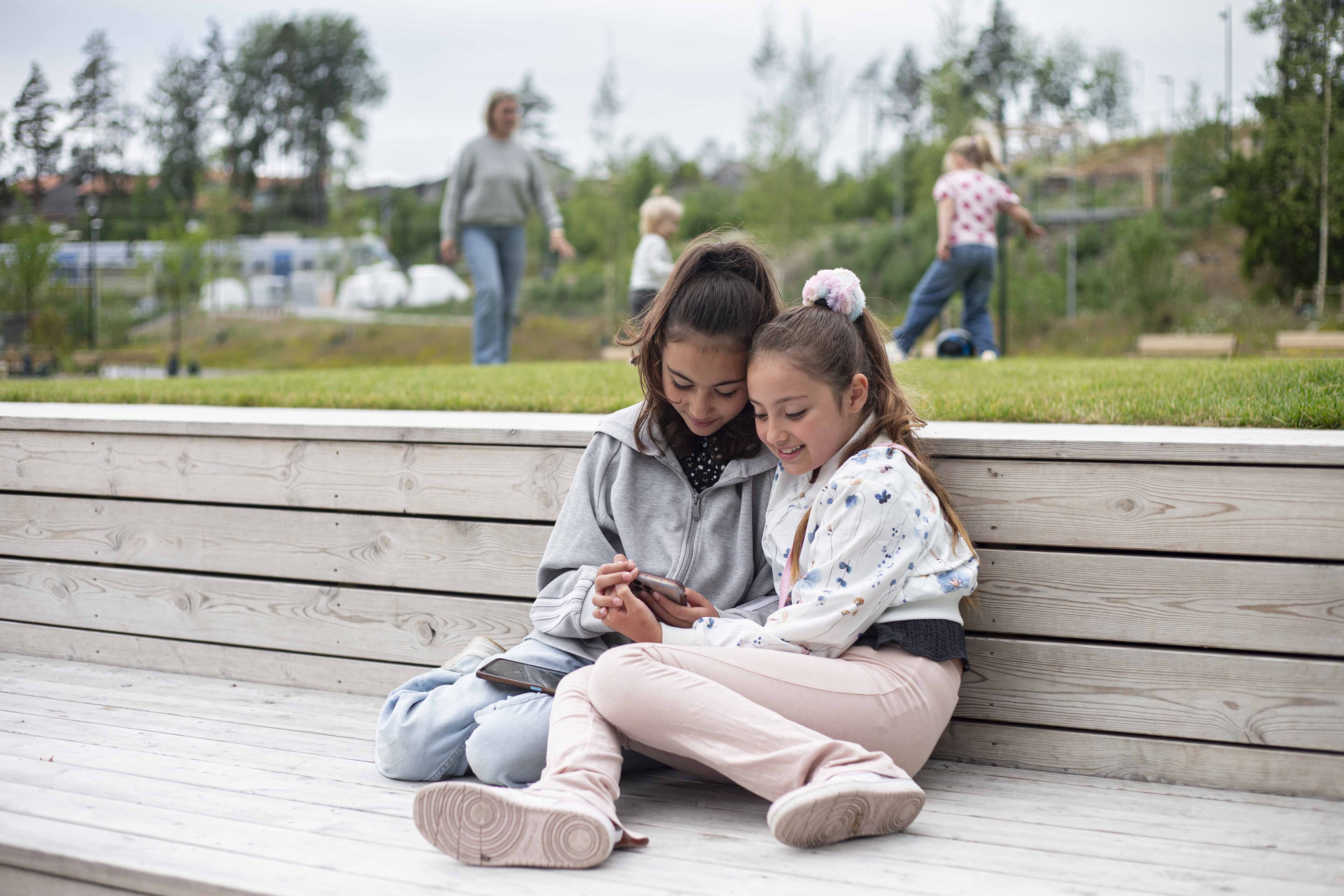 Två tjejer som tittar på telefonen i Vega med en familj som spelar fotboll i bakgrunden