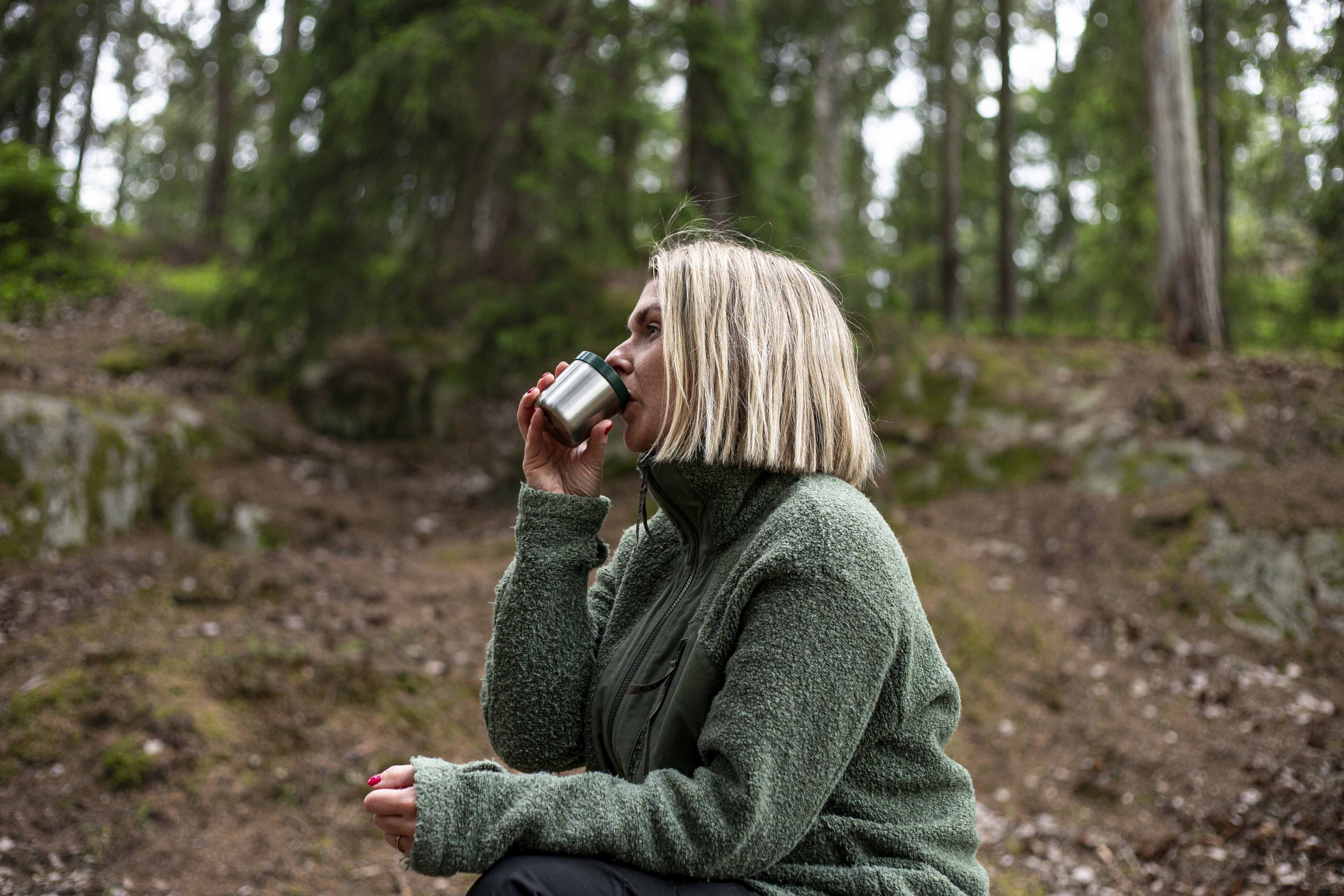 Kvinna i grön jacka dricker kaffe ur termoskopp i skogen
