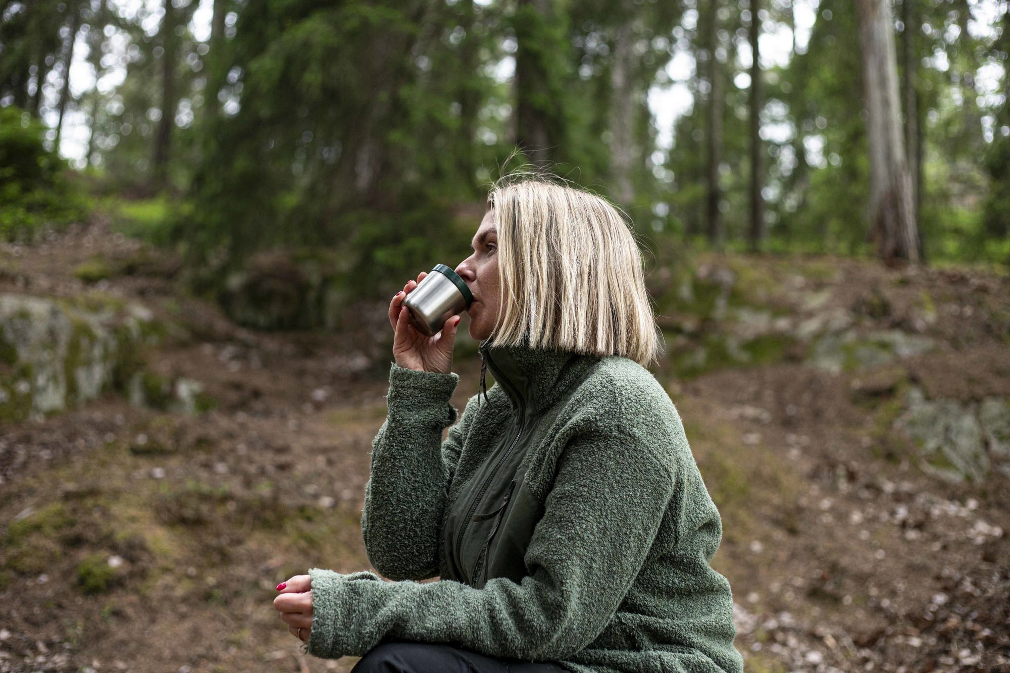 Kvinna i grön jacka dricker kaffe ur termoskopp i skogen