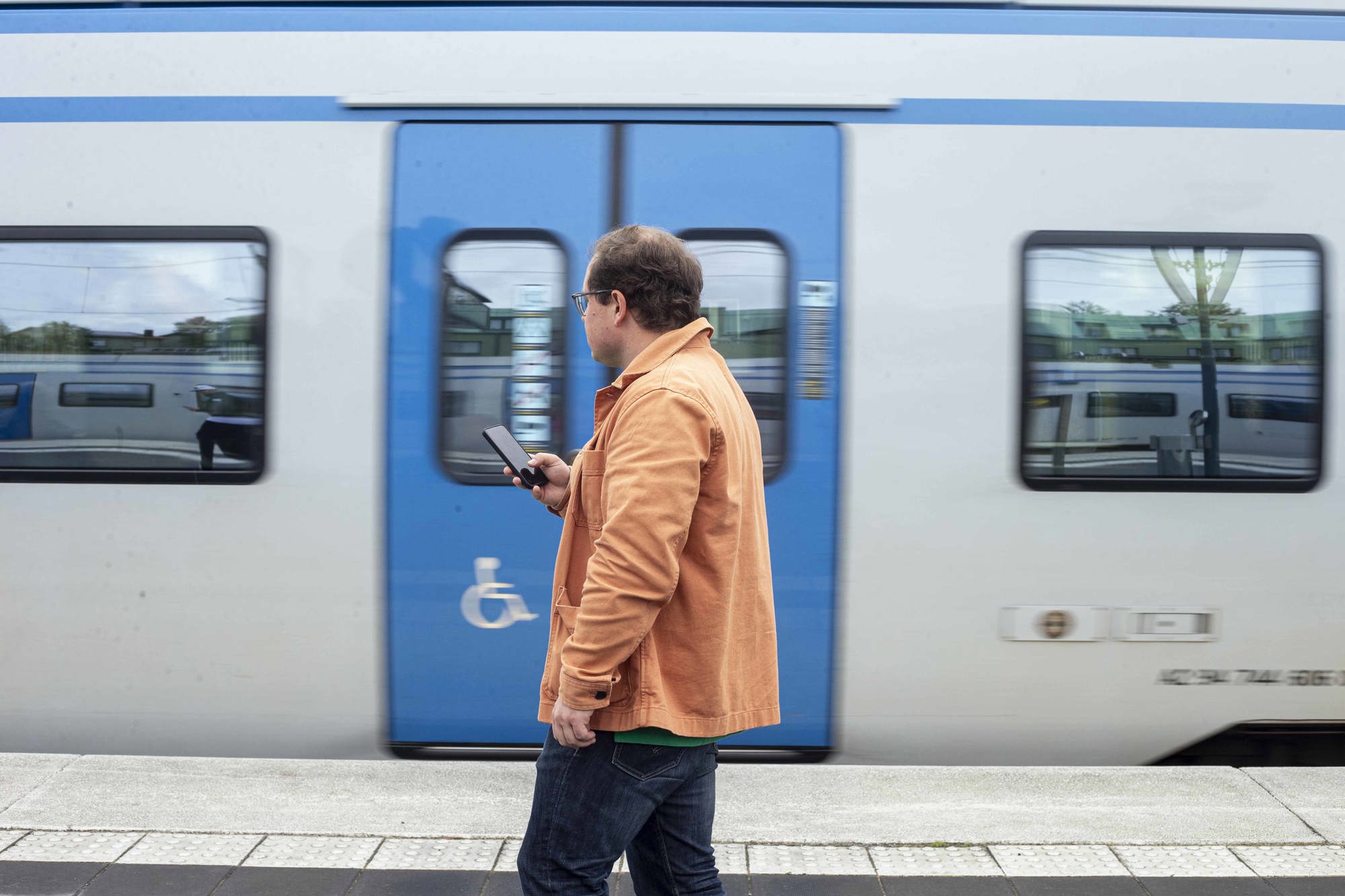 Man i orange jacka väntar på pendeln och kollar på telefonen