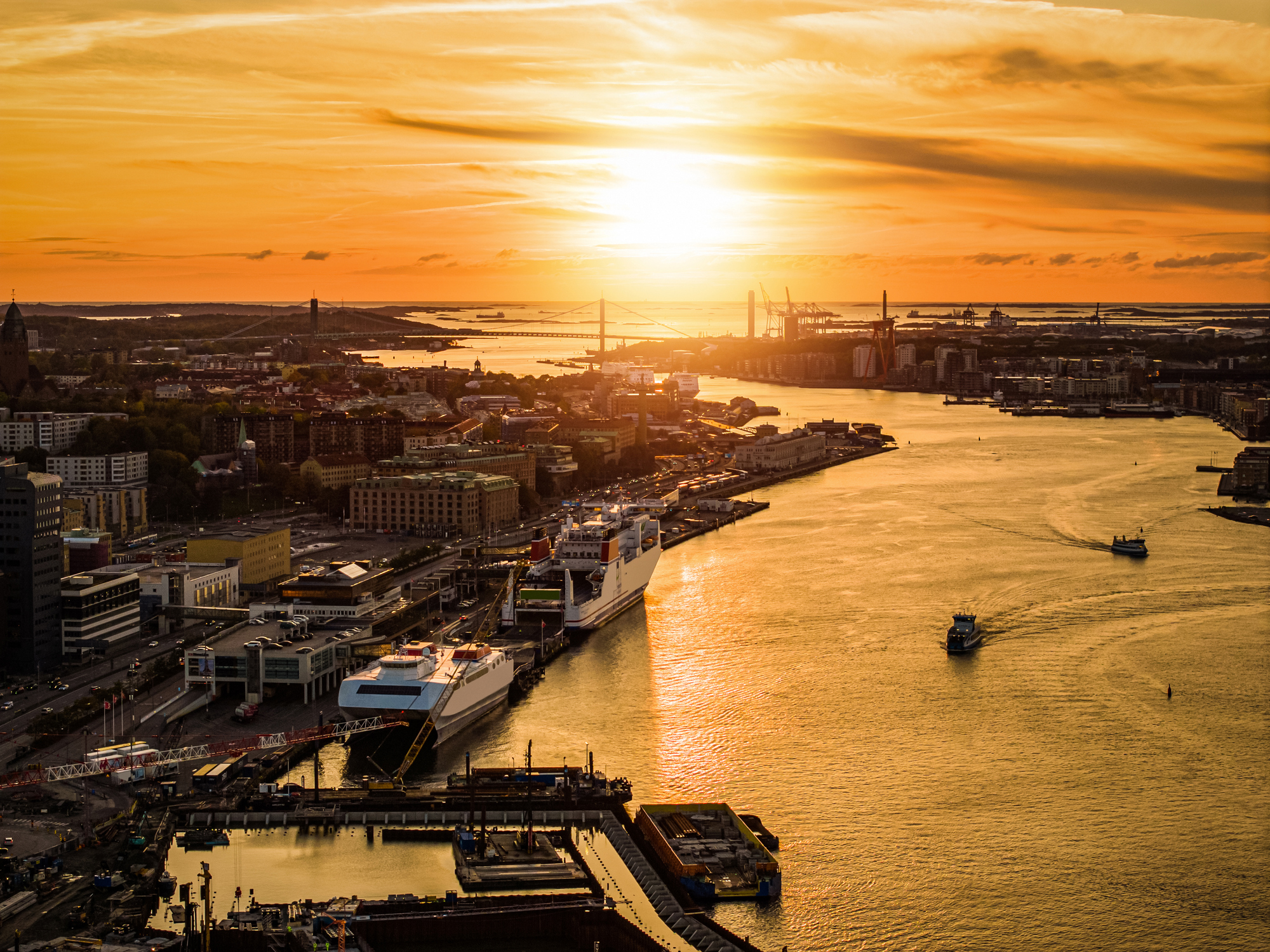 “Flygfoto över en stad med vatten och hamn i solnedgång, där den orange kvällssolen speglar sig i vattnet.”